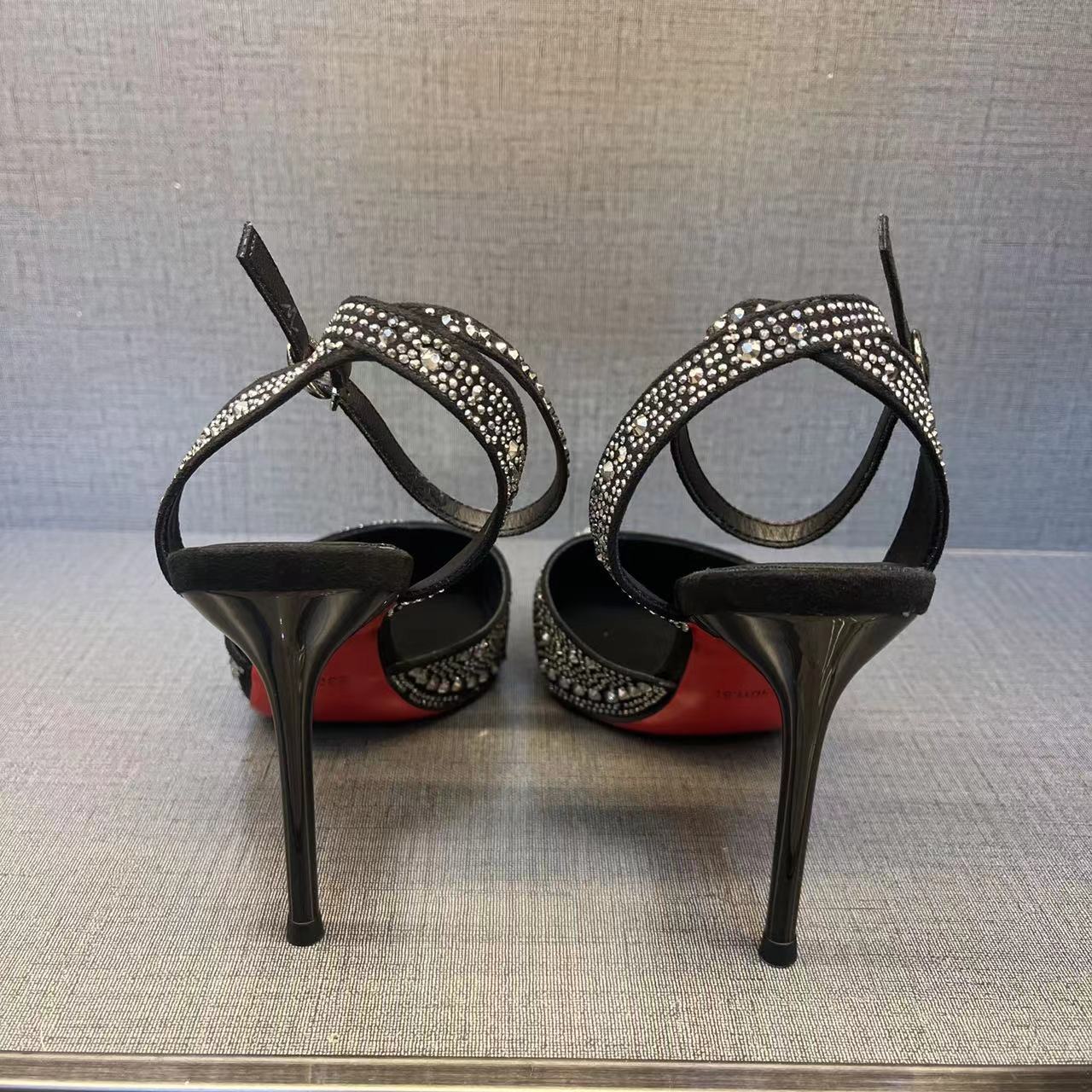 Black high-heeled shoes with red soles and rhinestone straps on a textured surface.