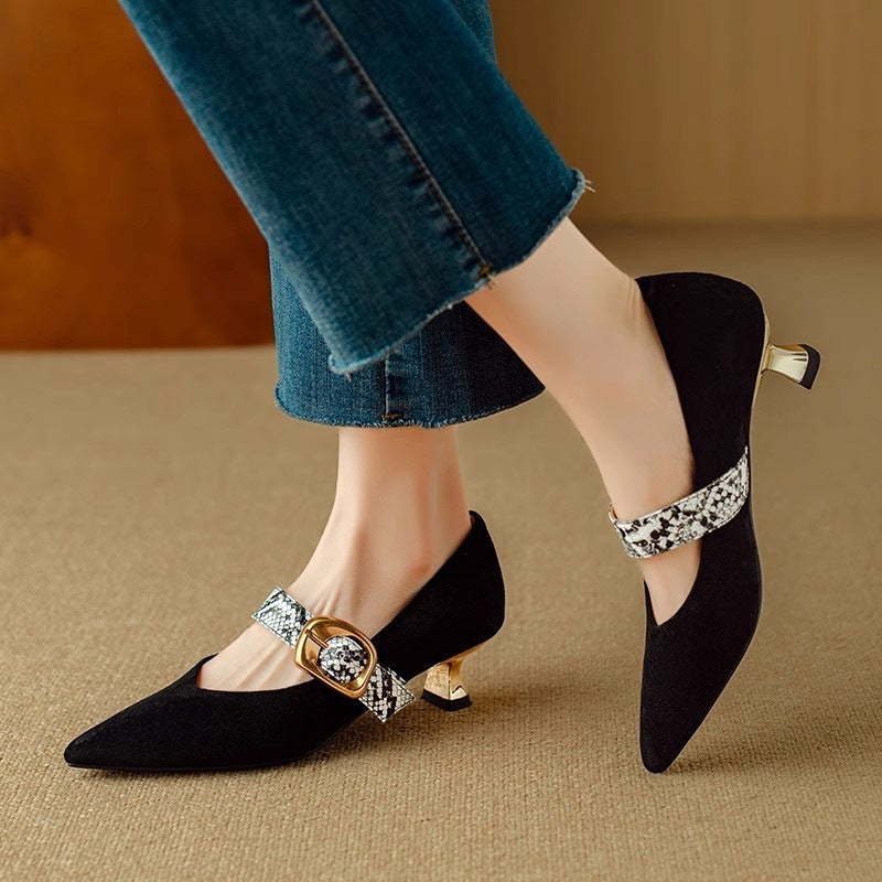 Black pointed-toe shoes with decorative buckles worn by a person in jeans on a beige surface.